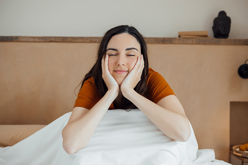 Dreamy attractive caucasian young woman sitting on comfortable bed in morning, smiling with closed eyes, resting chin on her hands. horizontal