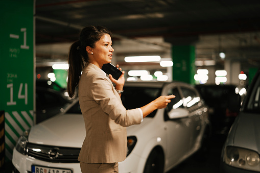 Businesswoman in suit unlocking car on parking. Beautiful woman talking to the phone.