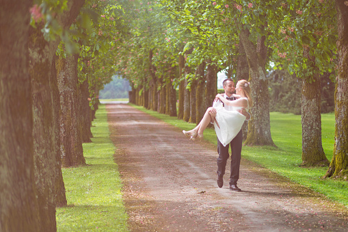 Young couple in wedding dress walking in nature forest path. Treelined path with man carrying his woman.