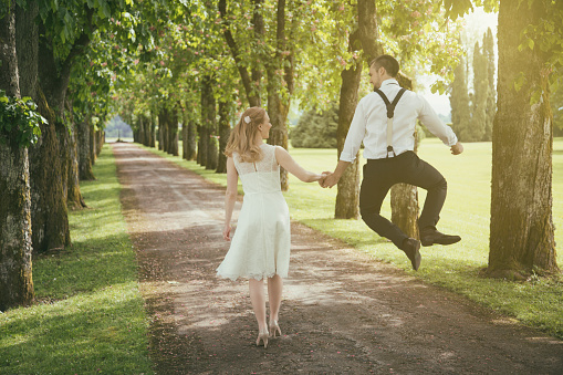 Young couple in wedding dress walking in nature forest path. Groom jumping in the air of joy. Walking from wedding.
