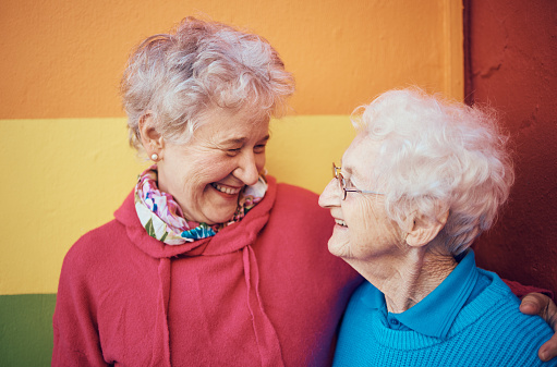 Friends, happy and retirement with a senior woman and friend outdoor together on a color wall background. Smile, freedom and glasses with mature female friendship bonding or laughing outside