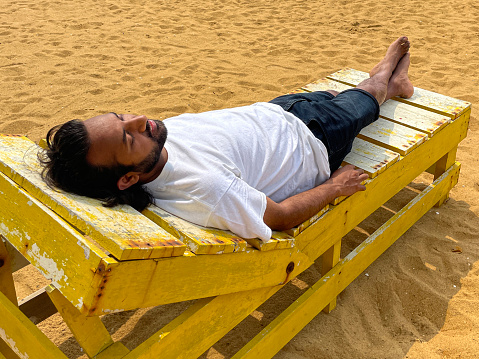Stock photo showing the golden sands of Mount Lavinia Beach, Colombo, Sri Lanka with wooden sun lounger being used by Indian man in front of al fresco dining area of rustic white painted tables and chairs background.