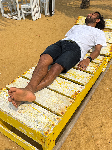 Stock photo showing the golden sands of Mount Lavinia Beach, Colombo, Sri Lanka with wooden sun lounger being used by Indian man in front of al fresco dining area of rustic white painted tables and chairs background.