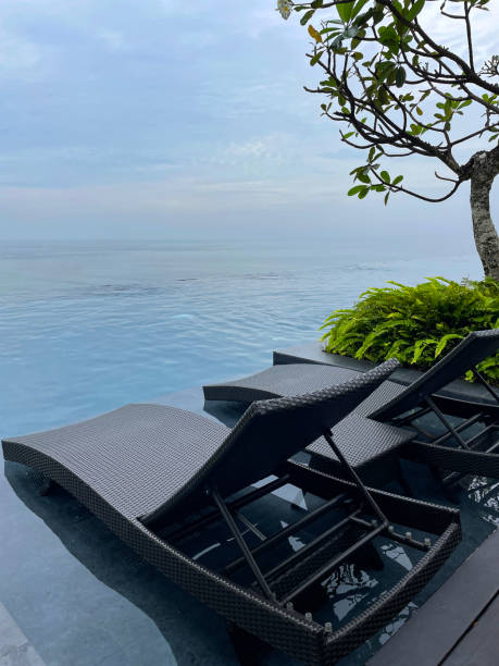 istock Close-up image of pair of poolside woven plastic sun lounger with side table besides infinity swimming pool, blue cloudy sky background, focus on foreground 1459322555
