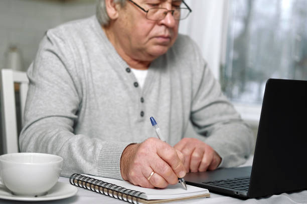 istock Senior man E-learning writing in notepad. Old male studying at home sitting at desk using laptop. Closeup hand writes in notebook with pen make note information. Older people distance learning concept 1459323162