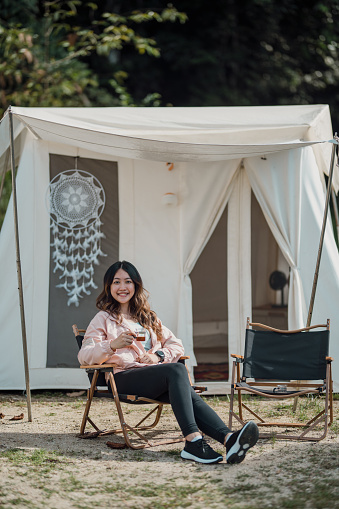 An Asian female sits relaxing outside a tent and enjoy a cup of coffee. Looking at camera. Outdoor activity, adventure travel, or holiday vacation concept.