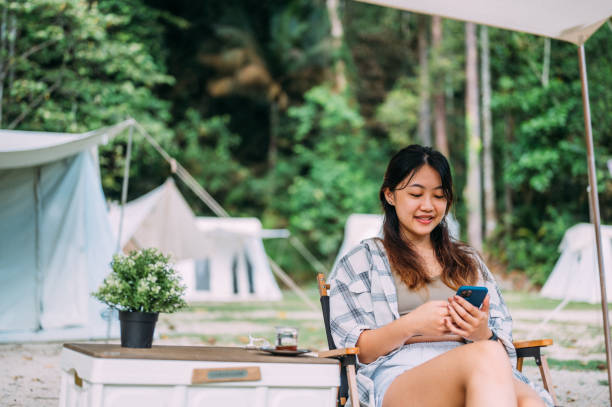 joven asiática sentada en su campamento y usando un teléfono inteligente en la naturaleza - oriental tent fotografías e imágenes de stock