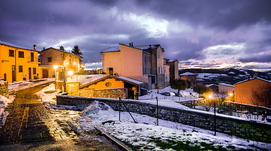 A winter atmosphere in the evening in the snowy stone alleys of the medieval village of Gualdo Tadino in Umbria, central Italy. The Umbria region, considered the green lung of Italy for its wooded mountains, is characterized by a perfect integration between nature and the presence of man, in a context of environmental sustainability and healthy life. In addition to its immense artistic and historical heritage, Umbria is famous for its food and wine production and for the high quality of the olive oil produced in these lands. Image in 16:9 ratio and high definition format.