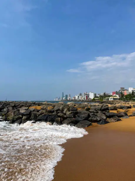 Photo of Image of rock groynes and seashore at Mount Lavinia beach in Colombo, Sri Lanka, sandy and rocky beach with sea and capital city visible in background, coastal erosion defences and blue sky on sunny day