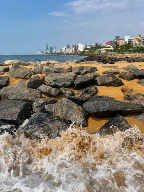 Photo of Image of rock armour / rip rap and waves splashing at Mount Lavinia beach in Colombo, Sri Lanka, sandy and rocky beach with sea and capital city visible in background, coastal erosion defences and blue sky on sunny day
