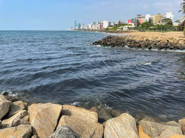 Photo of Image of view and groynes at Mount Lavinia beach in Colombo, Sri Lanka, sandy and rocky beach with, sea and capital city visible in background, coastal erosion defences and blue sky on sunny day