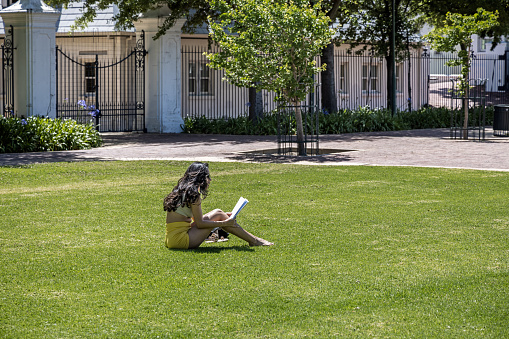 Cape Town, South Africa - December 10th 2022: Pretty young woman sitting on a green lawn in a public park while reading in a book
