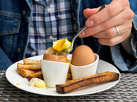 Stock photo showing close-up view of white crockery of tea plate and egg cups with white toast soldiers and two soft-boiled eggs one topped showing runny yellow yolk.