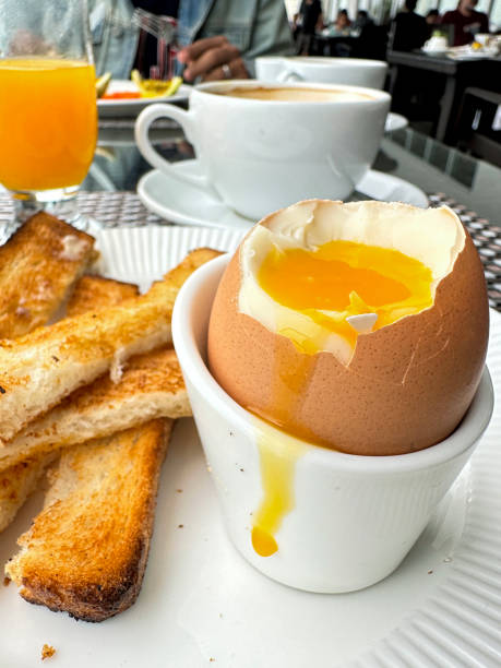 Close-up image of soft boiled egg with soldiers, runny orange yolk, from the cracked eggshell, white bread toast slices with butter, white eggcup, on white plate at cafe with coffee and orange juice in background, focus on foreground