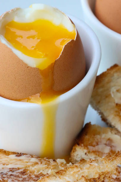 istock Full frame image of buttered toast soldiers with two soft boiled eggs in egg cups on white plate, topped soft boiled egg with runny yellow yolk, focus on foreground 1459329678