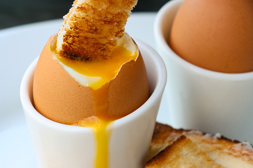 Stock photo showing close-up view of white crockery of tea plate and egg cups with white toast soldiers and two soft-boiled egg one topped showing runny yellow yolk.