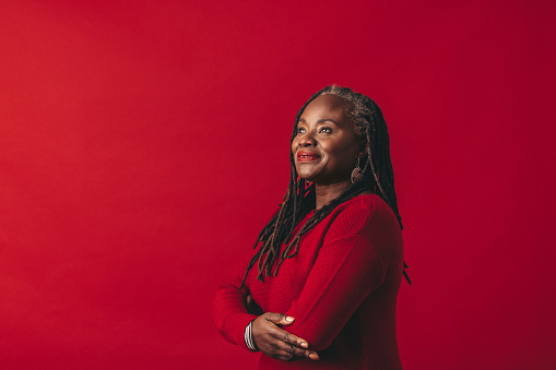 Pensive woman with dreadlocks looking away while standing against a red background. Mature black woman embracing her natural hair with confidence.