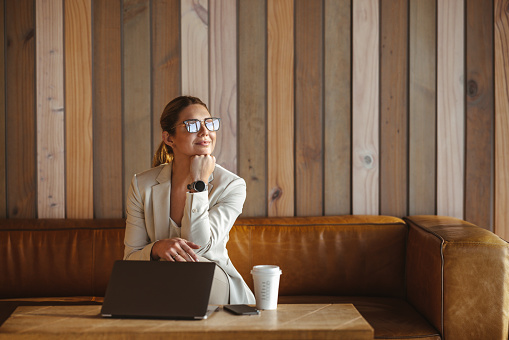 Thoughtful business woman sitting in a coffee shop with a laptop. Professional woman sitting on a couch in a cafe. Caucasian business woman working remotely.