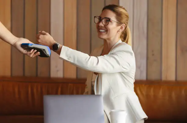 Woman scanning her smartwatch on a card machine to make a payment. Professional woman using NFC technology. Business woman buying coffee while working in a cafe.