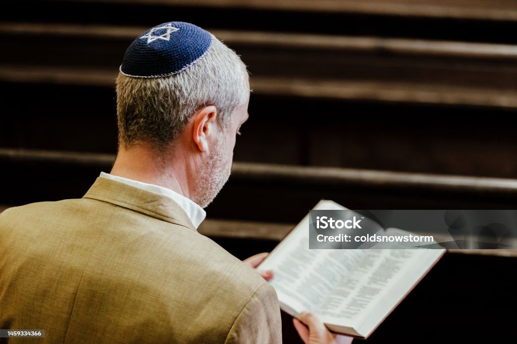 Jewish man wearing yarmulke while reading holy book in synagogue Portrait of a Jewish man reading a holy book of Jewish scriptures. The man is wearing a yarmulke on his head as he sits alone in the synagogue. Judaism Stock Photo