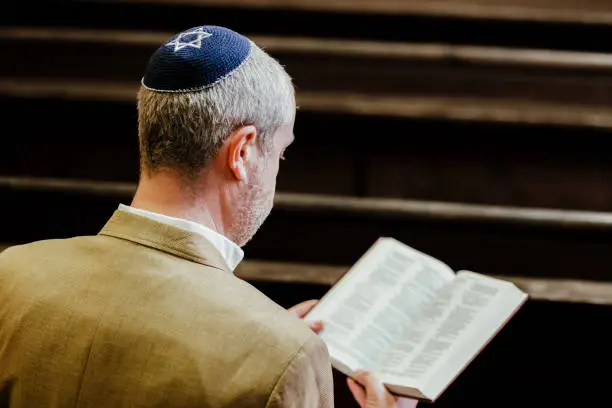 Portrait of a Jewish man reading a holy book of Jewish scriptures. The man is wearing a yarmulke on his head as he sits alone in the synagogue.
