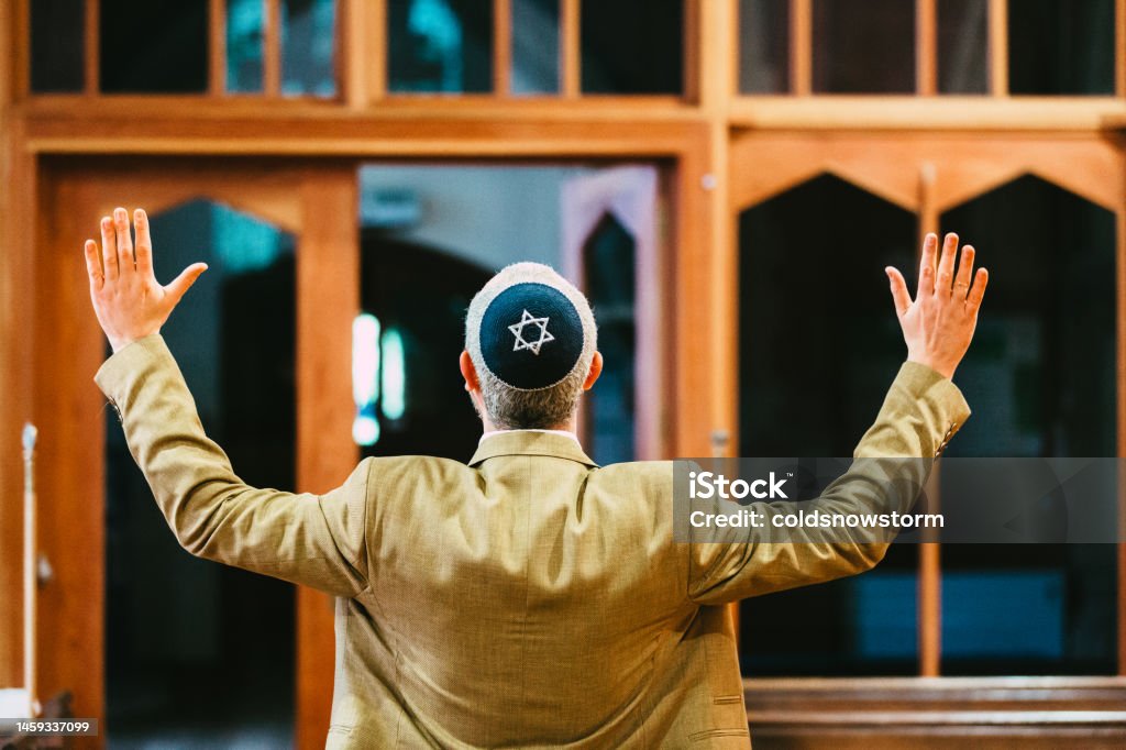 Celebrating Jewish man with arms outstretched in synagogue Rear view of a Jewish man celebrating in joy with his arms outstretched in the synagogue. He is wearing a yarmulke with the star of David embroidered on it. Judaism Stock Photo