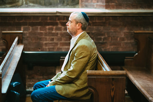 Portrait of a Jewish man sitting and praying in the synagogue. The man is wearing a yarmulke on his head as he sits alone in the synagogue.