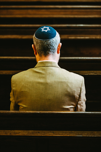 Portrait of a Jewish man sitting and praying in the synagogue. The man is wearing a yarmulke on his head as he sits alone in the synagogue.