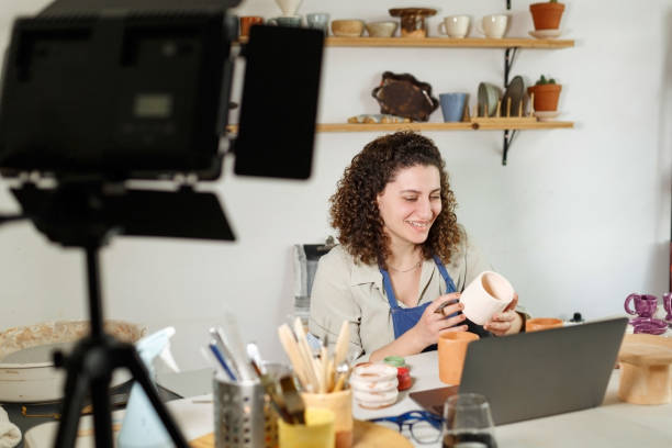 istock Females potter in blue apron making a vlog, recording online course, clay master class,lessons in her studion with earthenware shelf on background.Using led lamp,studio equipment 1459343564