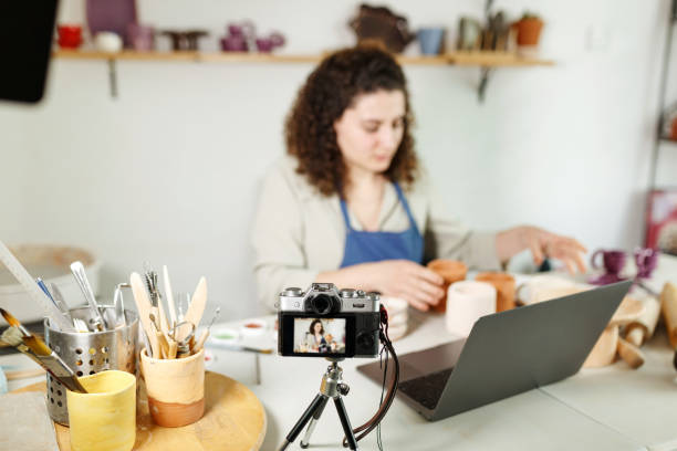 istock Females potter in blue apron making a vlog, recording online course, clay master class,lessons in her studion with earthenware shelf on background.Using led lamp,studio equipment 1459343587