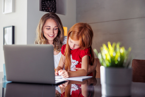 Mother holding daughter in her lap, tutoring her at home and helping with homework using laptop computer for online lessons; homeschooling and distance learning concept