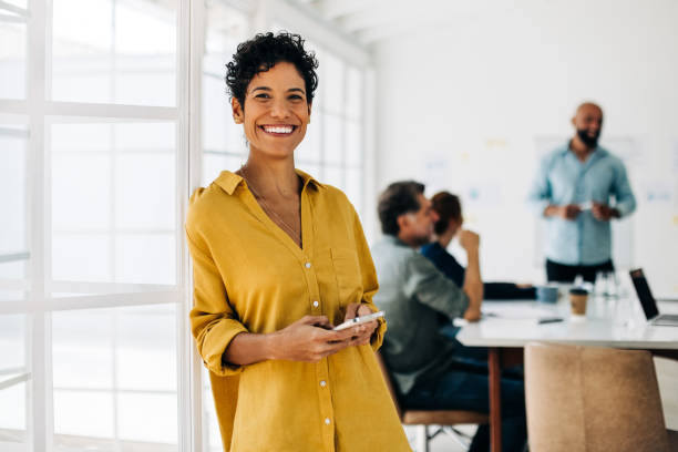 オフィスに立ち、携帯電話を使う女性プロフェッショナル - technology mobile phone clothing smiling ストックフォトと画像