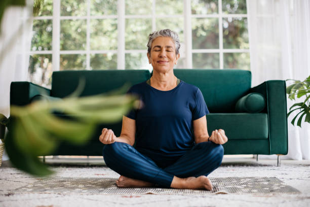 Relaxing the mind and finding inner peace with yoga: Senior woman meditating at home Senior woman meditating in lotus position at home, sitting on the floor in fitness clothing. Mature woman doing a breathing workout to achieve relaxation, peace and mindfulness. meditating stock pictures, royalty-free photos & images