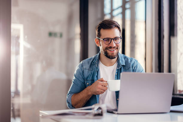 istock Handsome smiling young man sitting by window in office, drinking coffee while working on laptop. 1459348816
