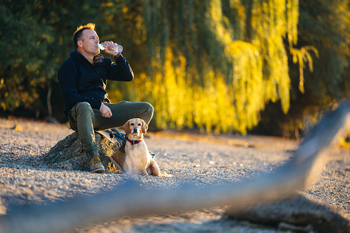 a good looking middle aged man having a sip of water by the river and the woods with his pet dog.