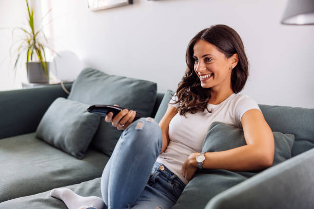 A smiling young woman sitting on the sofa and watching television. stock photo