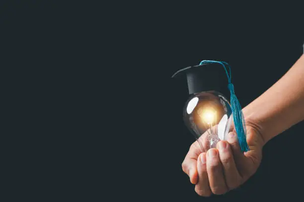 Photo of academy and success graduate education concept. businessman hand holding bright, electric light bulb with degree cap on black background. business education, knowledge, learning idea with copy space.