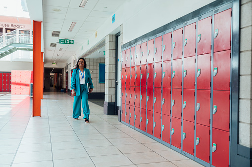 A wide angle view of a female head teacher walking through the hallways and lockers of her school. She is between meetings on her way to the next one carrying her notepad and her laptop. She is the head of a school in the North East of England.