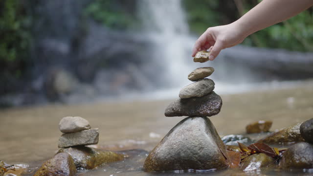 Hand stacking rocks nearby a waterfall in a forest.