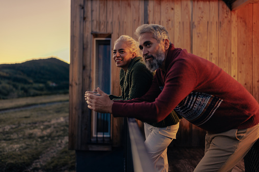 A senior Caucasian couple is in the mountains at their cabin, leaning against the railing on the patio, enjoying the scenic sunset view.