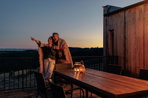 A senior couple is on their mountain cabin deck, pointing at the sky and gazing a the stars on a chilly autumn evening.