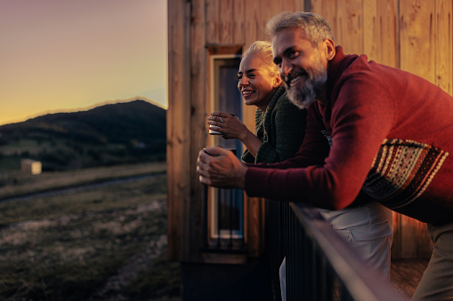 A mature Caucasian couple is in the mountains at their cabin, leaning against the railing on the patio, enjoying the sunset.