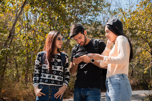 Multi-ethnicity group of young friends backpack traveling using technology smartphone finding map pointing direction, Caucasian and Asian tourist enjoy hiking ,walking, exploring in forest on vacation