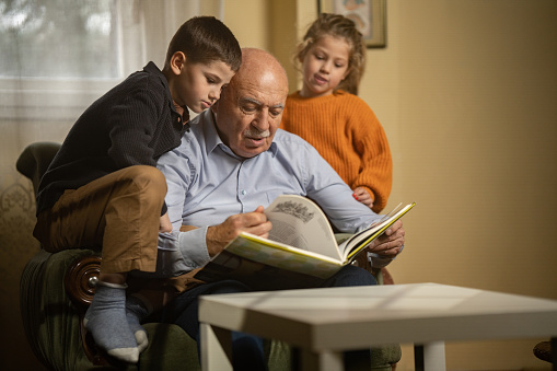 Old man reading stories to his young grandson and granddaughter in living room at home.