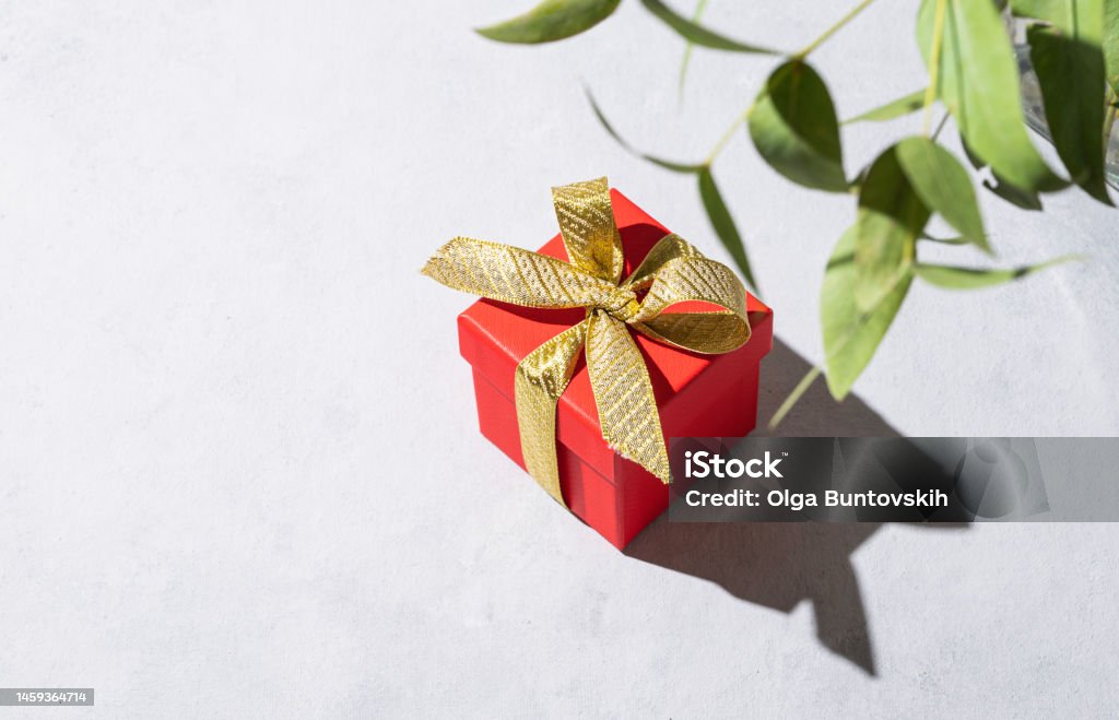 A red gift box with a gold ribbon on a gray background with shadow and branch flower. The concept of holiday photography. Surprise for Valentine's Day, birthday, wedding. A red gift box with a gold ribbon on a gray background with shadow and branch flower. The concept of holiday photography. Surprise for Valentine's Day, birthday, wedding. Copy space and top view. Abstract Stock Photo