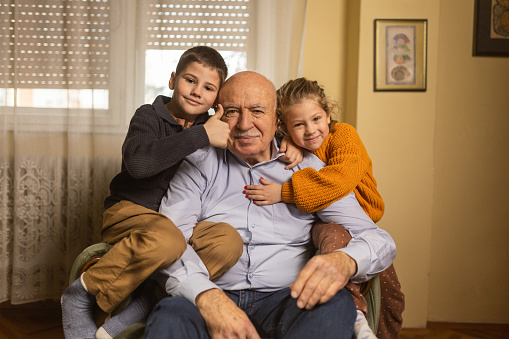 Old man sitting with his young grandson and granddaughter in living room at home.