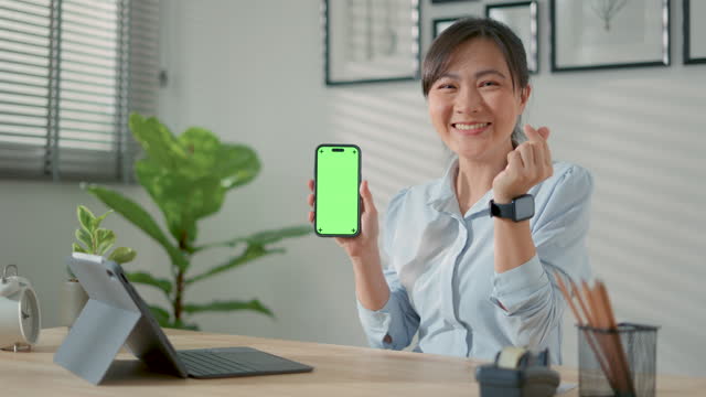 Asian woman happy smiling holding smart phone and showing green screen vertical orientation, make a mini heart by hand sitting at home office.