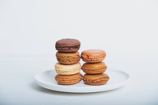 Stack of delicious freshly baked caramel, vanilla and chocolate flavoured macaron confections on white plate against white background. French sweet food speciality.