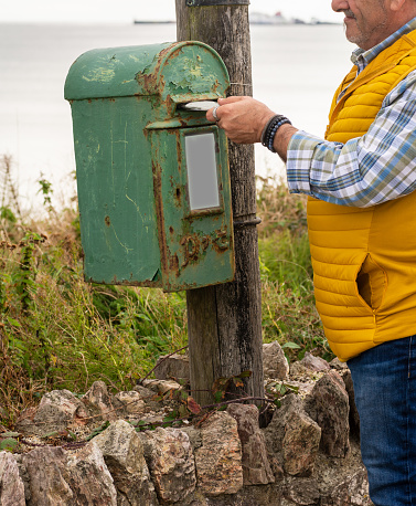 senior man putting letter in antique Irish mailbox, metal mailbox with rust, man in yellow vest. vertical image.