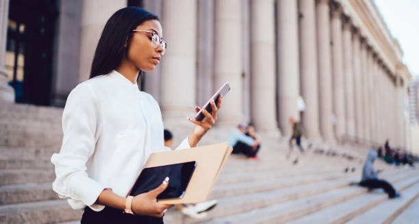 istock Long haired black woman in glasses browsing smartphone holding documents and tablet on stairs of building 1459381842